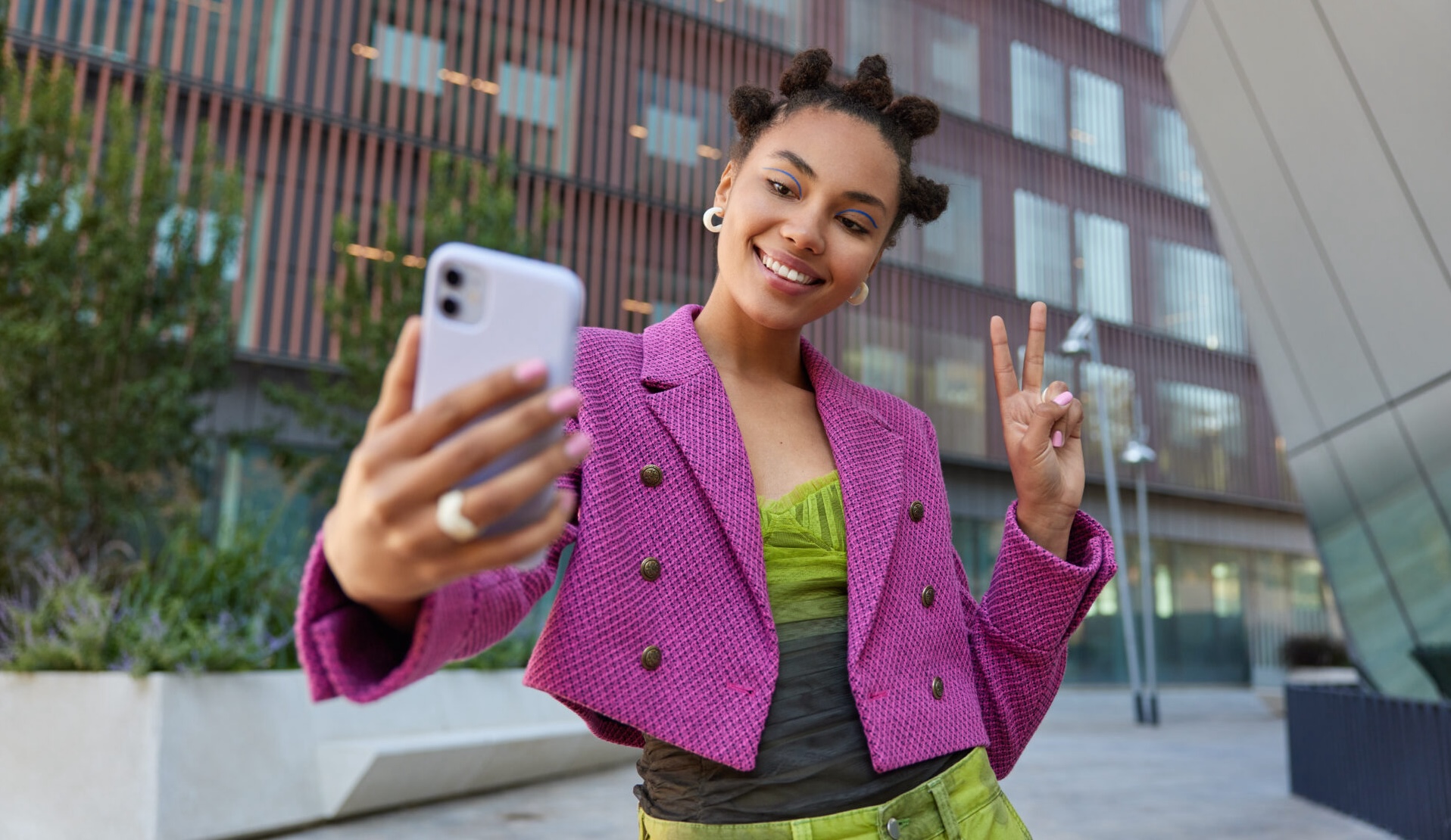 Attractive girl in fashionable outfit creats influence content shows peace sign at smartphone front camera poses for selfie smiles gladfully poses at urban place spends vacation at big city.