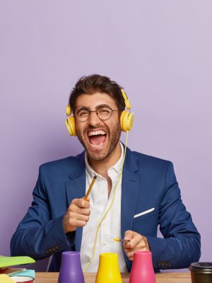 Positive finance expert has break, listens music in headphones, pretends playing drums on colorful cups with pencils, has overjoyed expression, dressed in formal clothes. Business and fun concept