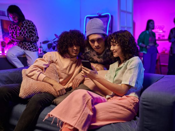 Three young friends in casualwear watching online videos in smartphone of cute brunette girl with curly hair while enjoying home party